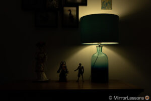 una muñeca japonesa y dos figuras de acción de final fantasy junto a una lámpara, en una cómoda.  La imagen está visiblemente subexpuesta.
