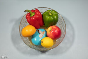 tazón de frutas y verduras colocado sobre una alfombra azul claro, que contiene una naranja, un pimiento rojo, un pimiento verde, una manzana, un limón y una piedra pintada de azul