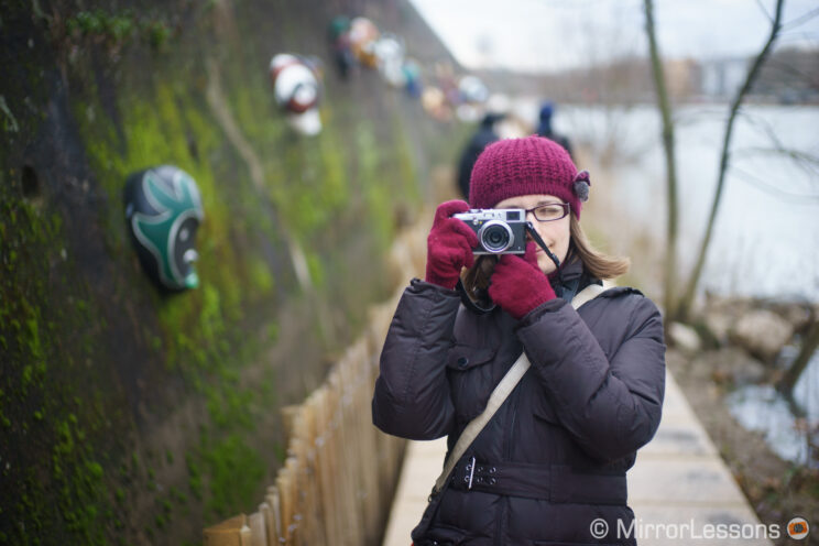 woman in winter clothes using a vintage camera along a modern river promenade